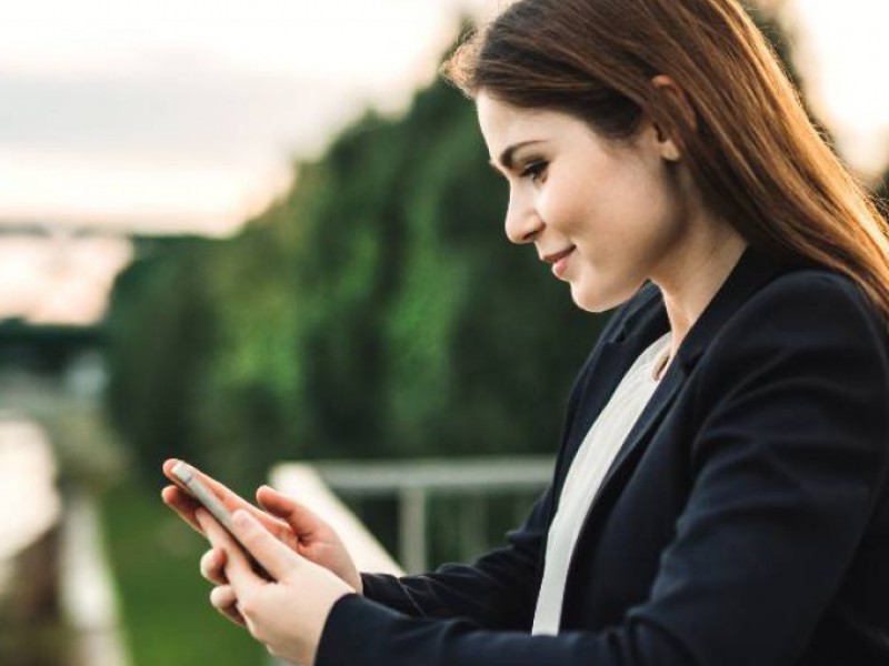 Jeune femme souriante au bord d'un fleuve faisant une simulation sur son smartphone.